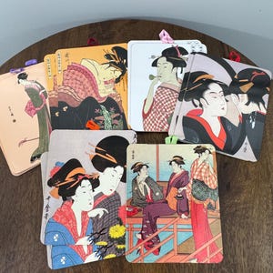 May include: A collection of square coasters featuring Japanese Ukiyo-e art prints. Each coaster showcases a different scene with women in traditional attire, using a variety of colors including red, black, and gold. The coasters have a glossy finish and are arranged on a wooden surface.