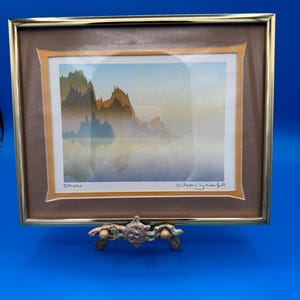 May include: A framed watercolor print titled DAWN, featuring a misty landscape with silhouetted mountains reflected in calm water. The artwork is encased in a thin gold-colored metal frame with a brown mat and a decorative gold-toned inner border.