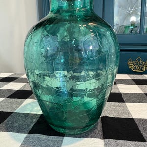 May include: A large, teal-colored glass vase with a crackled texture. The vase has a rounded body and a narrow neck. The glass is transparent, allowing light to pass through. The vase is sitting on a black and white checkered tablecloth.