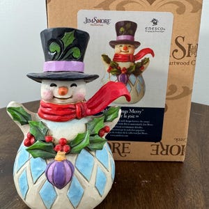 May include: A Jim Shore snowman figurine with a black top hat, red scarf, and holly accents. The snowman has a smiling face and a patterned body with a purple ornament. The figurine is displayed with its original packaging.