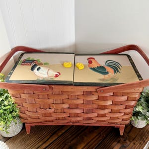 May include: A brown wicker picnic basket with a hinged lid featuring painted farm scenes. The lid depicts a rooster, hen, and chicks. The basket has a red handle and legs, perfect for outdoor dining.