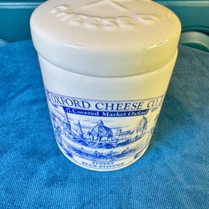 May include: A cream-colored ceramic crock with a matching lid featuring embossed text reading CHEESE CO. The side displays a blue printed illustration of Oxford city architecture with the text OXFORD CHEESE CO. 17. Covered Market Oxford.