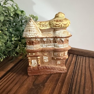 May include: A ceramic house-shaped figurine featuring a brown and cream color palette with a textured roof, multiple windows, and a front door. The structure has a multi-level design with detailed architectural elements and a weathered finish.