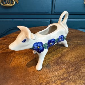 May include: A white ceramic creamer in the shape of a cow, adorned with blue floral patterns. The creamer has a handle and a spout, and is standing on four legs. The cow's face has a small blue eye.