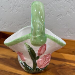 May include: A ceramic basket with a green handle and rim, and a white interior. The exterior features pink tulip designs with green leaves. The basket is set on a wooden surface.