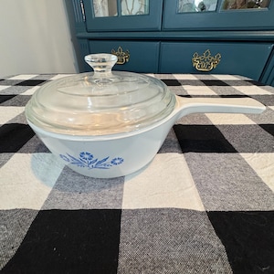 May include: White casserole dish with a clear glass lid and a long white handle. The dish has a blue floral design around the rim. The dish is sitting on a black and white checkered tablecloth.