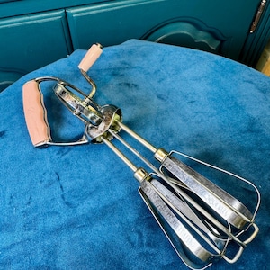 May include: Vintage hand-crank egg beater with a metal frame and blades. The handles are a light pink color. The beater is resting on a blue velvet surface. A classic kitchen tool.