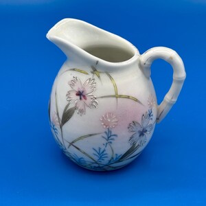 May include: A white ceramic creamer pitcher featuring a delicate hand-painted floral pattern with pink, blue, and green botanical motifs. The pitcher has a rounded body, a wide pouring spout, and a handle textured to resemble a bamboo stalk.