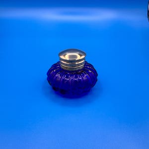 May include: A small, round, cobalt blue glass inkwell with a ribbed, fluted texture and a polished, reflective silver-toned metal cap. The container has a wide, squat base that tapers slightly toward the top, featuring a smooth, rounded lid.