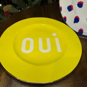 以下が含まれることがあります： 白い文字で「oui」と書かれた明るい黄色のプレート。プレートにはシルバーの縁があります。青と赤のドット模様の別のプレートが背景に部分的に見えます。装飾的なプレートです。