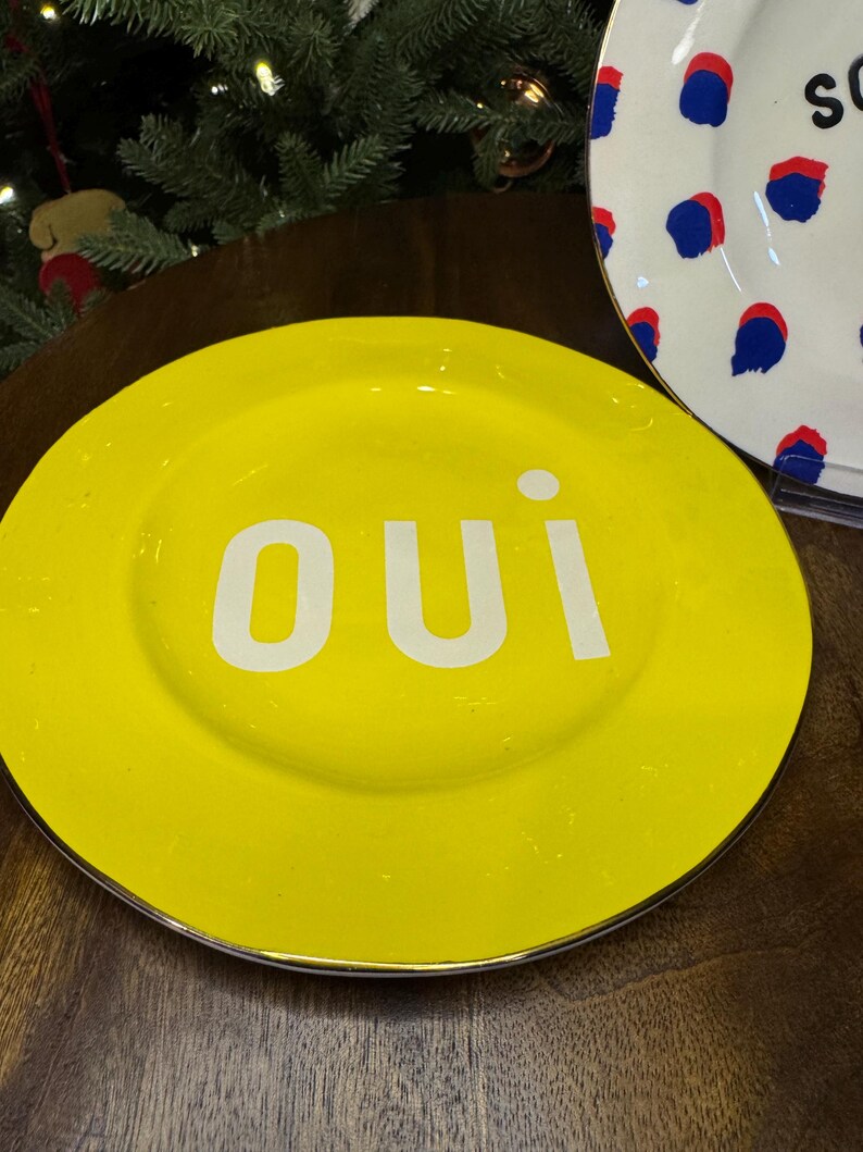 以下が含まれることがあります： 白い文字で「oui」と書かれた明るい黄色のプレート。プレートにはシルバーの縁があります。青と赤のドット模様の別のプレートが背景に部分的に見えます。装飾的なプレートです。