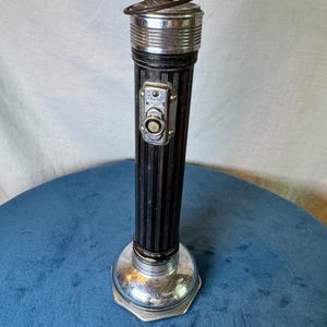 May include: A vintage cylindrical flashlight featuring a black body with vertical pinstripes, a polished chrome head, and a matching chrome base. The device includes a rectangular metal switch plate with a central button and a small metal hanging loop.