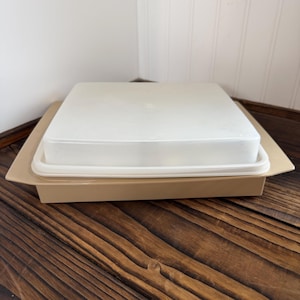 May include: A tan square container with a clear, square lid. The container sits on a wooden surface. The lid is slightly curved and has a small handle in the center. The container is made of plastic.