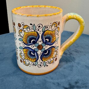 May include: A ceramic mug with a white base, adorned with a symmetrical floral pattern in shades of blue, yellow, and brown. The rim and handle are yellow, with a decorative yellow trim. The mug sits on a blue surface.