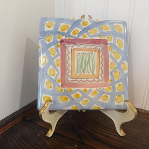 May include: A square ceramic tile featuring a central illustration of green stems with small pink flowers inside a scalloped border. The background displays a blue lattice pattern with yellow dots, all framed by a thick, muted red square border.