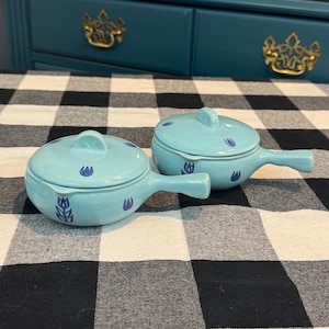 May include: Two light blue ceramic soup bowls with lids and handles. Each bowl features a blue floral design and a matching lid with a small handle. The bowls are placed on a black and white checkered tablecloth.