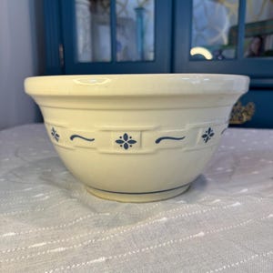 May include: Cream-colored ceramic mixing bowl with blue floral and scroll design. The bowl has a wide rim and a slightly flared shape. A blue line accents the base of the bowl. A classic kitchenware item.