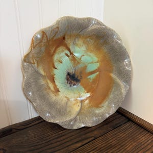 May include: A round ceramic bowl with a scalloped, wavy edge. The glaze features a blend of earthy brown, soft sage green, and cream tones, with a dark, textured center. The surface has a glossy finish with visible radial brushstroke patterns.