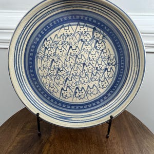 May include: A round ceramic plate with a blue and white design. The center features a pattern of numerous cat faces. The rim has blue stripes and a dotted border. The plate is displayed on a black metal stand.