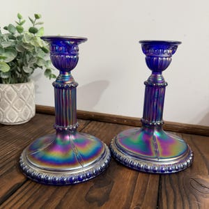 May include: Two iridescent blue glass candle holders. The candle holders have a round base, a fluted column, and a small cup to hold the candle. The glass has a rainbow sheen. Decorative home decor.