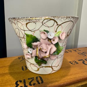 May include: Cream-colored ceramic vase with pink roses and green leaves. The vase has a gold-trimmed rim and gold accents. Pink and gold swirls decorate the exterior. A decorative floral container.
