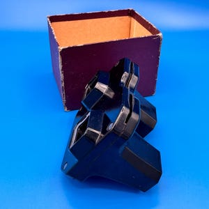 May include: A black plastic stereoscope viewer with two eye-pieces and a central slot for viewing slides, positioned in front of its original square, dark purple cardboard box with a tan interior lining, set against a solid blue background.