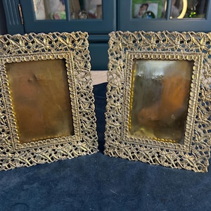 May include: Two ornate, vintage picture frames. The frames have a gold-colored, filigree design with a rectangular opening. The frames are made of metal and have a decorative floral pattern around the edges. The frames are placed on a dark blue surface.