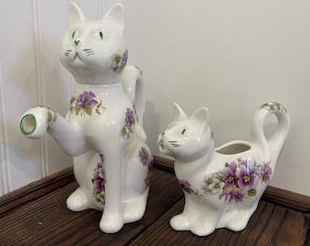 May include: A white ceramic cat-shaped teapot and creamer set. The teapot has a handle and a spout with a green rim. Both pieces are decorated with purple and white floral patterns and green eyes.