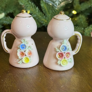 May include: A pair of pink ceramic salt and pepper shakers. Each shaker has a bulbous body, a handle, and a gold-colored lid. The shakers are decorated with colorful floral designs.