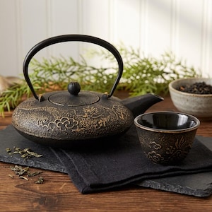 May include: A black cast iron teapot with a gold-colored dragon and cloud relief pattern, featuring a curved handle and a matching small cup. The teapot has a round lid with a central knob, and the cup displays a similar textured design.