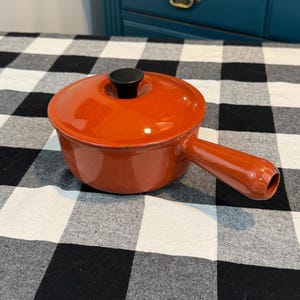 May include: A small, orange enameled cast iron pot with a lid and a single handle. The lid has a black knob. The pot is sitting on a black and white checkered surface. The pot is designed for cooking.