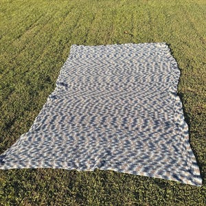May include: A rectangular crochet blanket featuring a variegated pattern of blue, white, and tan yarn. The textile displays a consistent, repeating stitch texture throughout its surface, creating a dense and uniform appearance across the entire piece.
