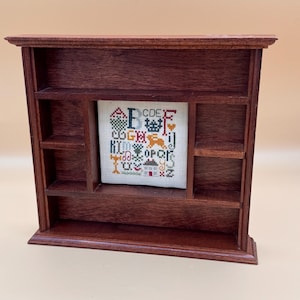 May include: A dark brown wooden display shelf with multiple compartments featuring a square cross-stitch sampler in the center. The sampler displays the alphabet in various colors, including letters A through Z, alongside small motifs like a house.