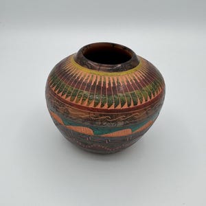 May include: A round, squat ceramic pot featuring a dark brown rim and a body decorated with horizontal bands of etched patterns. The design includes alternating sections of terracotta, green, and black, with geometric shapes and wavy lines.