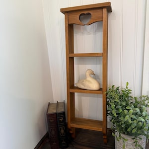 May include: A tall, narrow, three-tiered wooden wall shelf with a medium brown finish. The top section features a decorative cutout in the shape of a heart. A light-colored wooden duck figurine is positioned on the middle shelf of the unit.