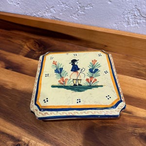 May include: Square ceramic tile with a hand-painted design. The tile features a figure in a hat and traditional clothing, surrounded by floral elements in blue, orange, and green. The border is blue and orange.