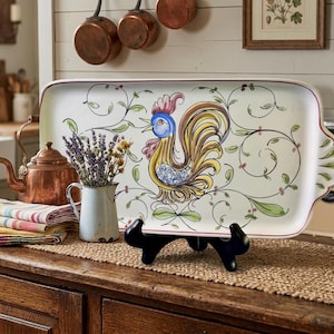 May include: A rectangular ceramic serving tray featuring a hand-painted rooster with blue, yellow, and red plumage, surrounded by delicate green vine motifs on a cream background. The tray has a scalloped edge and rests on a black display stand.