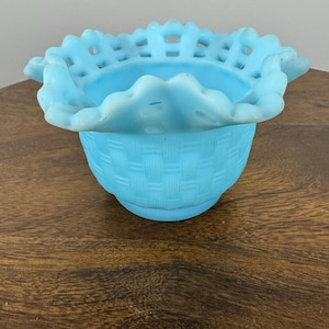 May include: A light blue, vintage-style glass basket with a woven texture on the body and a ruffled, openwork rim. The basket is made of opaque glass and sits on a wooden surface. This decorative piece is likely used for display or storage.