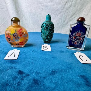 May include: Three decorative snuff bottles labeled A, B, and C. Bottle A is round and amber-colored with floral patterns. Bottle B is a teal, intricately carved vessel. Bottle C is a rectangular clear glass bottle with a blue insert featuring a floral design.