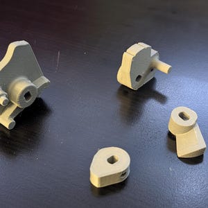 Peut inclure: Quatre pièces mécaniques imprimées en 3D, de couleur gris clair, sont disposées sur une surface sombre. Les pièces comprennent deux grands composants aux formes géométriques, une petite projection cylindrique et deux petites pièces avec des trous et des ouvertures carrées. Les pièces semblent être des composants d'un mécanisme plus large.