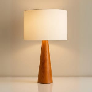 Puede incluir: Una lámpara de mesa de madera con base cónica y pantalla blanca. La base es de madera marrón claro y la pantalla es cilíndrica. La lámpara está sobre una superficie blanca y el fondo es de color neutro.