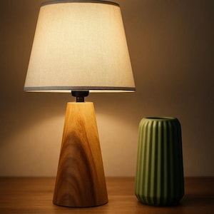 May include: A wooden table lamp with a beige lampshade and a light grey trim. The lamp has a wooden base with a natural wood grain. A green, ribbed vase sits beside the lamp.