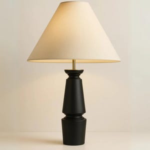 Puede incluir: Una lámpara de mesa negra con una pantalla beige. La lámpara tiene un tallo color latón y una base cilíndrica escalonada. La pantalla tiene forma de cono. La lámpara está diseñada para uso en interiores.