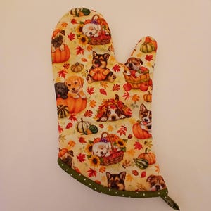 May include: A green and white oven mitt with a fall-themed design featuring adorable puppies and pumpkins. The mitt has a polka dot trim.