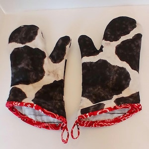 May include: A pair of oven mitts with a cowhide pattern in black and white. The cuffs are trimmed with a red paisley fabric. Each mitt has a loop for hanging.
