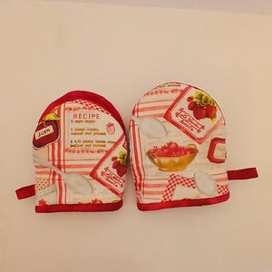 May include: A pair of oven mitts with a red and white strawberry design. The mitts have a red trim and a loop for hanging. One mitt features a recipe for jam. The other mitt has a bowl of strawberries.