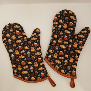 May include: A pair of black oven mitts with a brown trim. The mitts feature a repeating pattern of fall-themed treats, including pie slices, cupcakes, and mugs of hot chocolate.