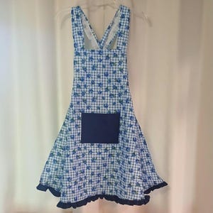 May include: A blue and white gingham apron with a floral pattern and a navy blue pocket. The apron has a ruffled hem and adjustable straps, perfect for cooking or crafting.