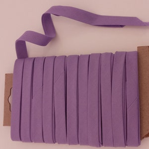 May include: A roll of lavender purple fabric trim, folded and stacked on a brown cardboard backing. The trim is approximately 1/2 inch wide.