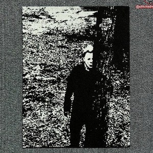 May include: Black and white image of a person wearing a mask, standing in front of a tree. The image is grainy and has a vintage look.